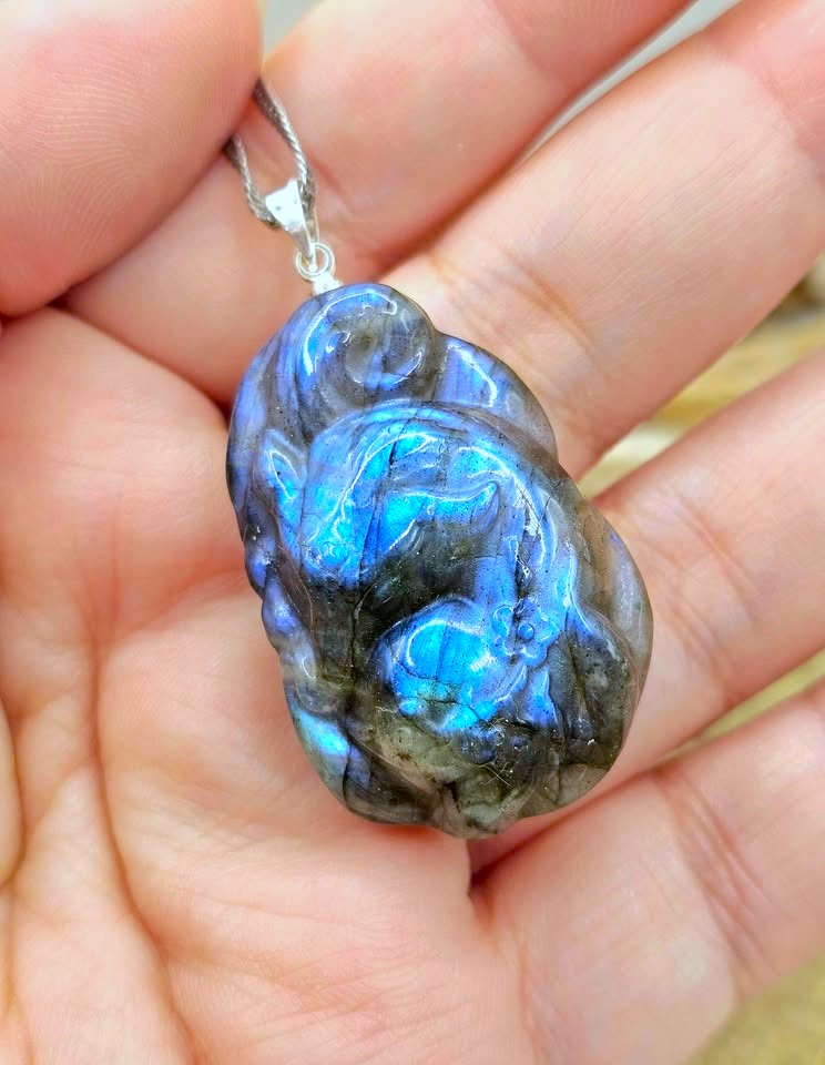 A polished labradorite pendant with a blue and green iridescent flash, held in a person's hand. The pendant is set in a silver-toned metal loop with a silver chain.