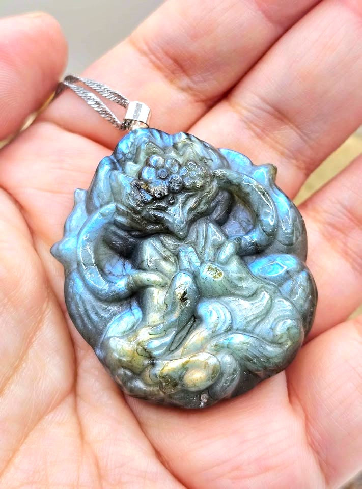 A hand-carved labradorite pendant featuring a detailed nine tailed fox goddess design, held in a person's palm to show its size. The pendant displays natural labradorite coloration with blue, green, and gray tones, suspended from a silver chain.