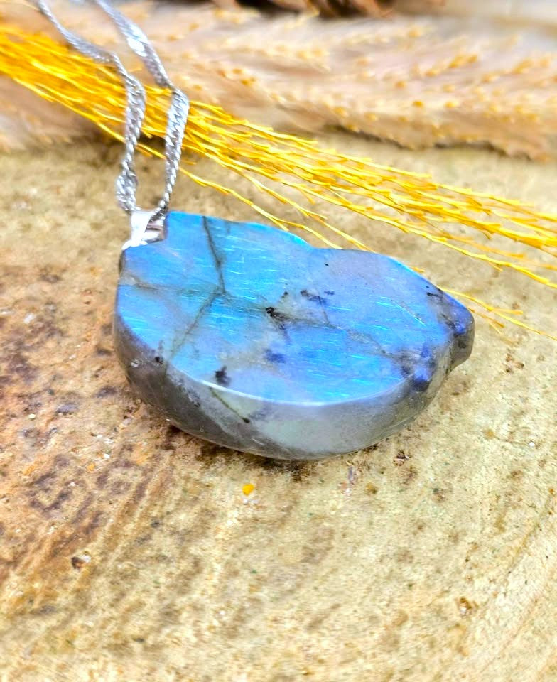 A teardrop-shaped labradorite gemstone pendant necklace with a silver chain, resting on a textured wood surface with dried yellow grass.