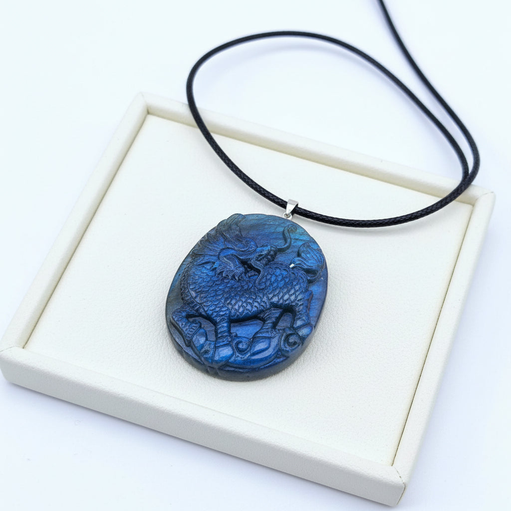 A carved blue stone pendant featuring a detailed  foo dog motif, suspended from a black cord with a silver-toned bail, displayed on a textured tan surface with dried grass and a pinecone in the background.
