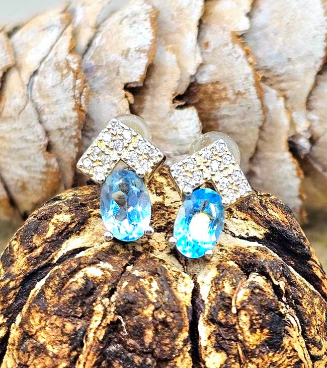 A pair of drop earrings with oval light blue gemstones and square silver settings covered in small clear stones, displayed on a textured brown wood surface with a blurred natural background.