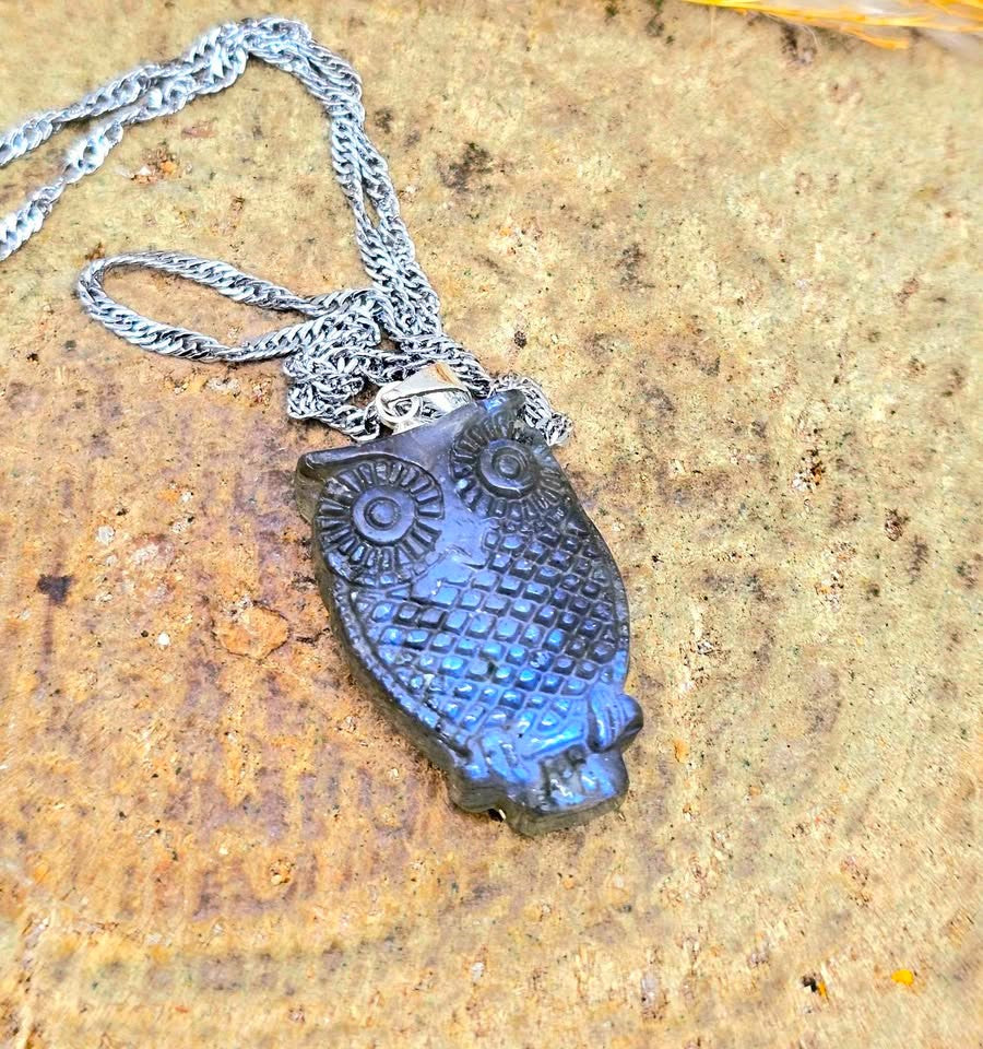 A carved owl pendant necklace made of dark blue iridescent stone, hanging from a silver-toned chain on a textured tan stone surface.