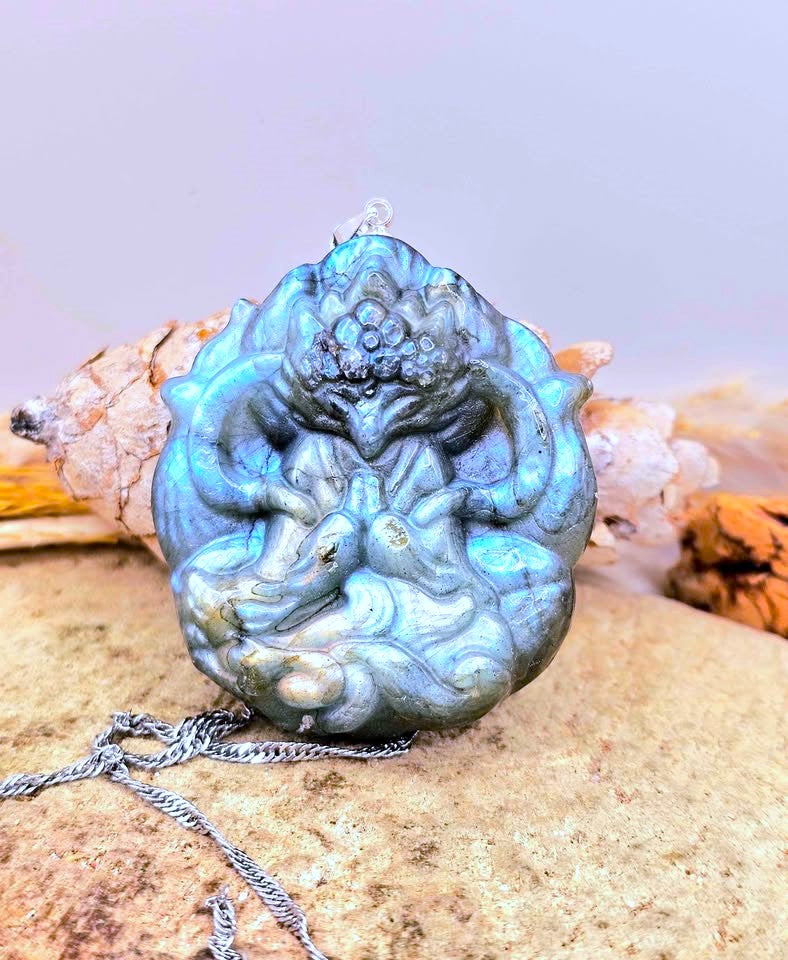 A carved nine tailed fox goddess pendant or charm with a metallic blue-green iridescent finish, featuring a stylized figure design, resting on a sandy surface with driftwood and a silver chain.