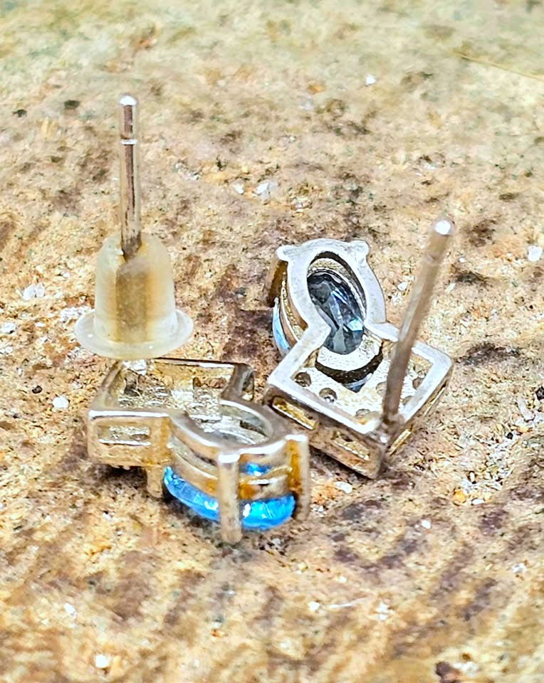 A pair of silver-toned stud earrings with oval blue gemstones, shown disassembled with the posts removed and standing upright on a textured sandy surface.