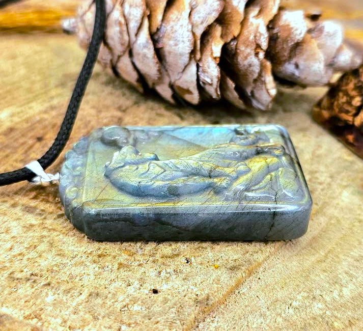 A rectangular carved labradorite pendant necklace with a raised figure design, resting on a textured wood surface with a pinecone in the background.