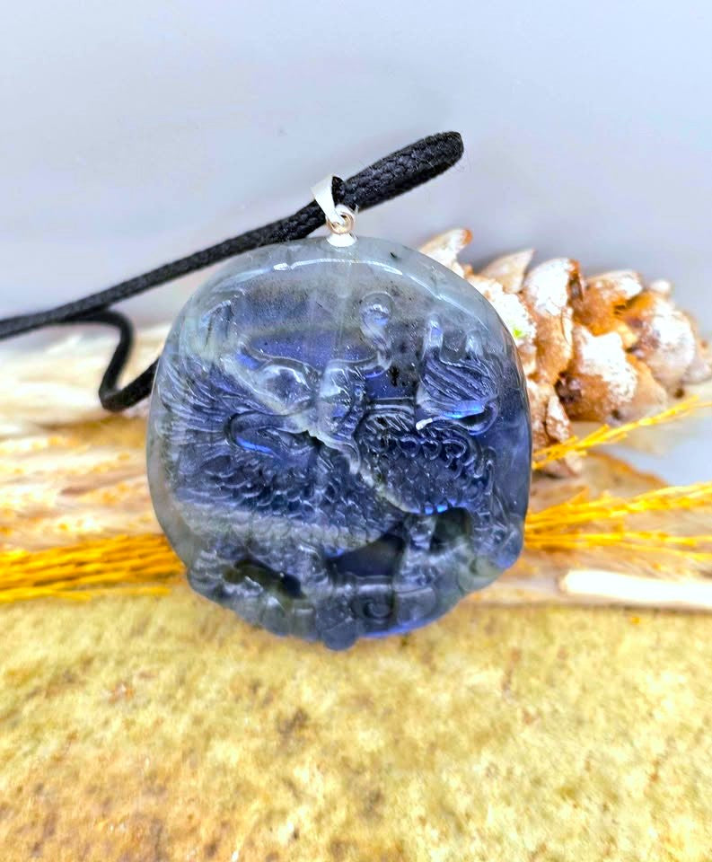 A carved blue glass pendant featuring a detailed foo dog design, suspended from a black cord with a silver-colored metal loop, displayed against a neutral background with decorative dried grass and shells.