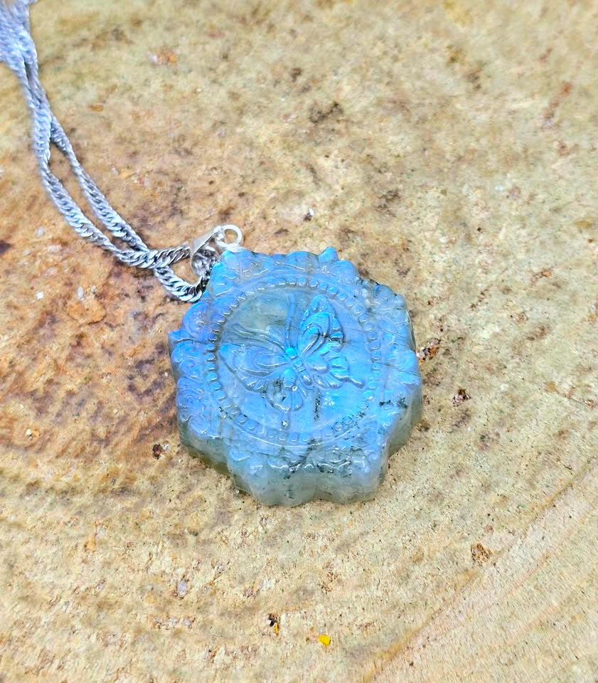 A blue-green labradorite pendant necklace with a silver chain, resting on a textured sandy surface.