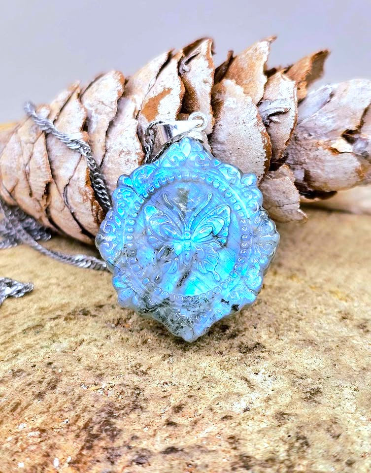 A silver-toned necklace with a round, textured turquoise pendant featuring an embossed butterfly design, styled on a rock with a pine cone.