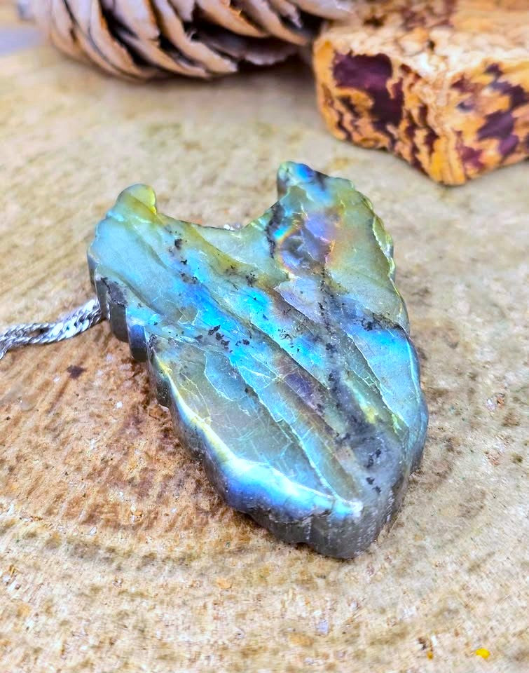 A raw labradorite stone pendant with a silver chain, resting on a textured stone surface with pinecones and wood in the blurred background.
