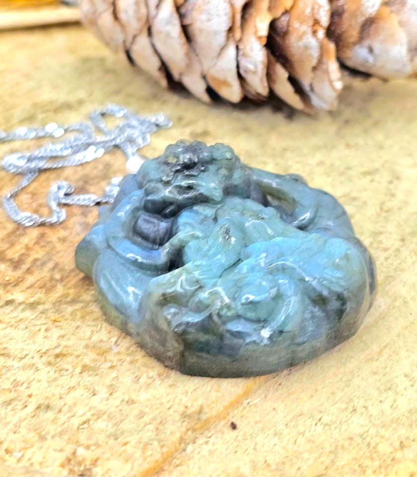 A carved jadeite dragon pendant in shades of green and blue, shown on a silver chain against a textured beige stone background with a pinecone prop.