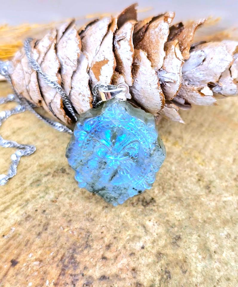 A blue labradorite crystal pendant necklace with a silver-toned chain, displayed on a textured stone surface next to a dried pine cone.