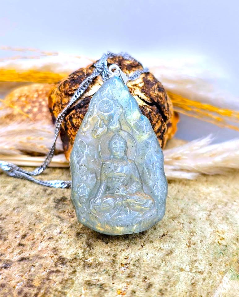 A teardrop-shaped pendant carved with a seated Buddha figure, made from a pale blue-gray stone with subtle gold flecks, hanging from a silver chain. The pendant is styled on a textured stone surface with dried grass and a brown shell in the background.