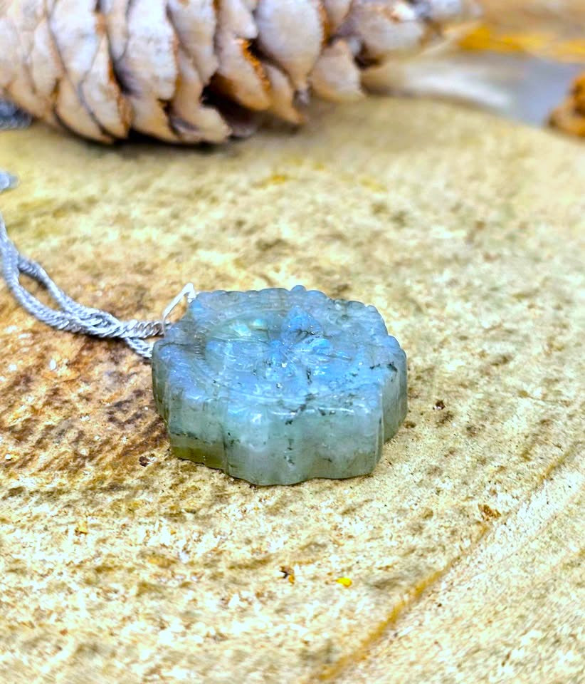 A raw, faceted Labradorite gemstone pendant on a silver cord, resting on a textured stone surface with a pinecone in the background.