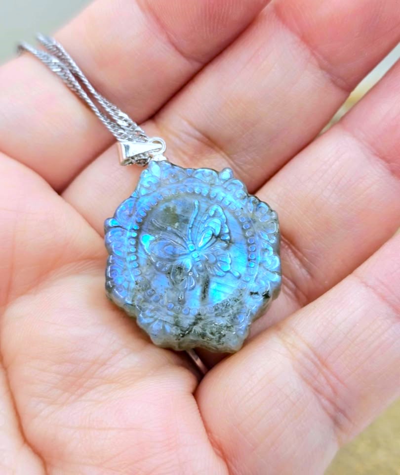 A close-up of a hand holding a silver pendant necklace featuring a highly iridescent, blue-green labradorite stone with a floral relief design.