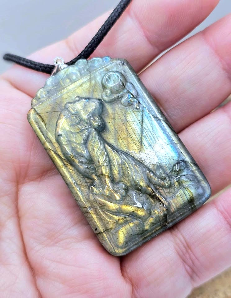 A rectangular labradorite pendant necklace with a raised cat design, held in a person's hand. The pendant displays a metallic gold and silver shimmer with natural labradorite play-of-color, suspended from a black cord.