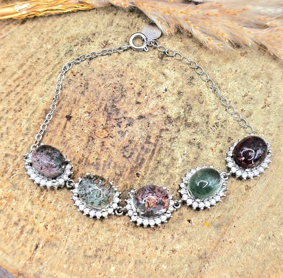 A silver chain bracelet featuring five oval gemstone cabochons in silver bezel settings, displayed on a textured wood slice background with dried grass.