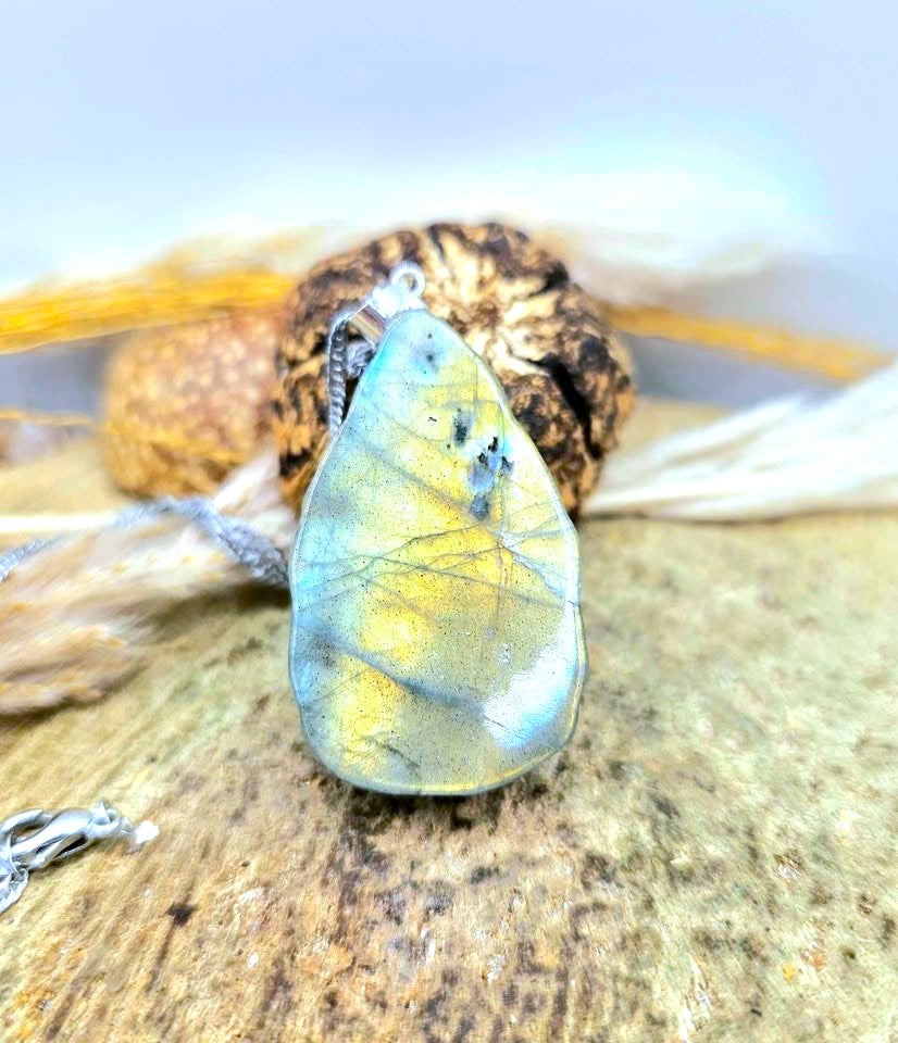 A teardrop-shaped labradorite pendant with a silver bail, resting on a textured stone surface with dried grass and a small silver clasp visible.