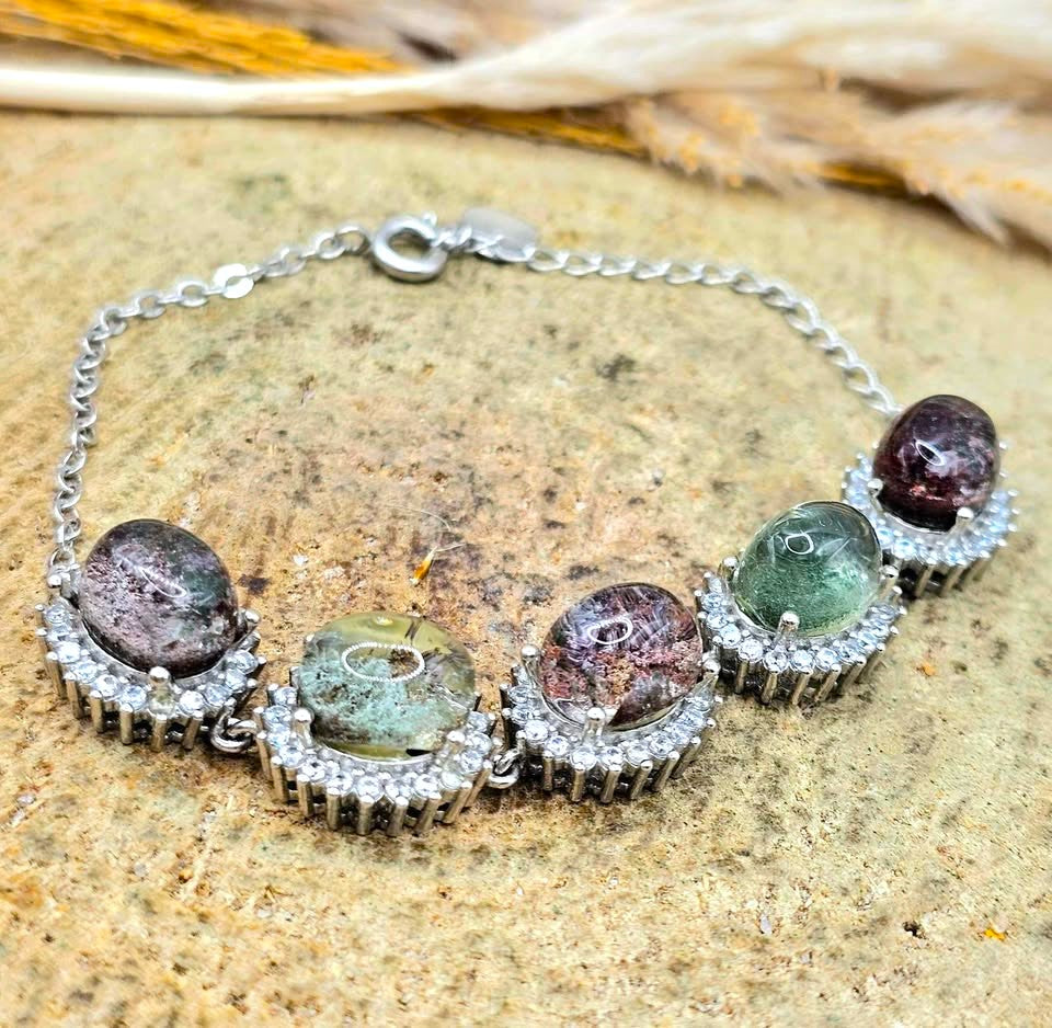 A silver chain bracelet featuring five oval gemstone cabochons in silver bezel settings, displayed on a textured stone surface.