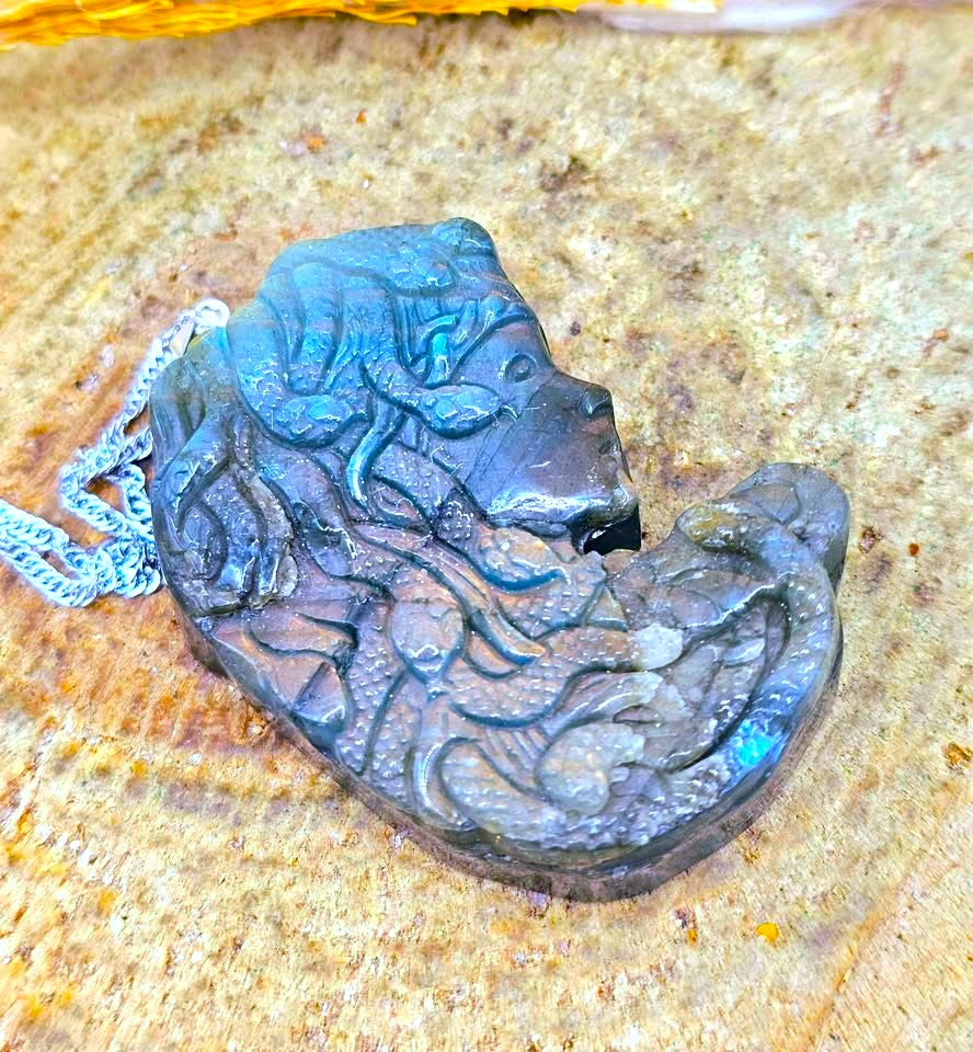 A carved dark stone pendant with a stylized face and flowing hair details, attached to a silver chain, resting on a textured tan stone surface.