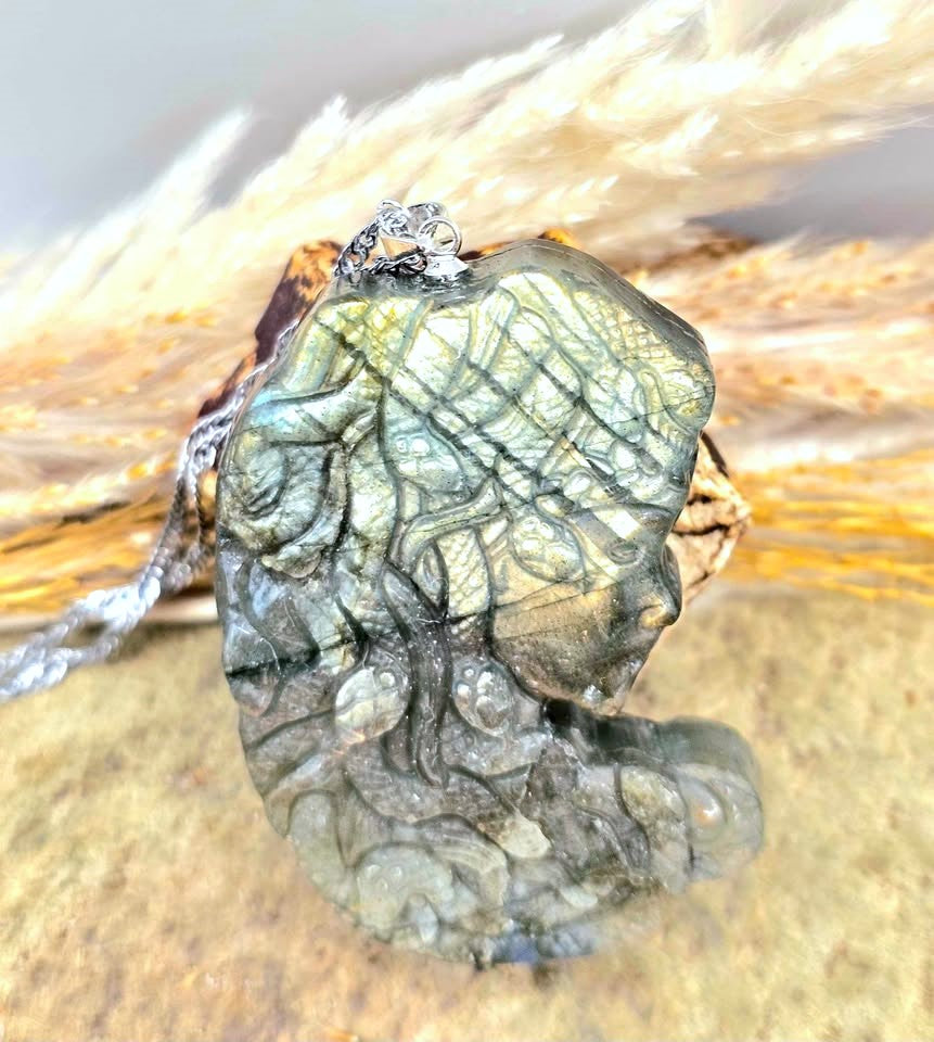 A close-up of a carved labradorite stone pendant with a silver bail, resting on a textured surface with dried pampas grass in the background.
