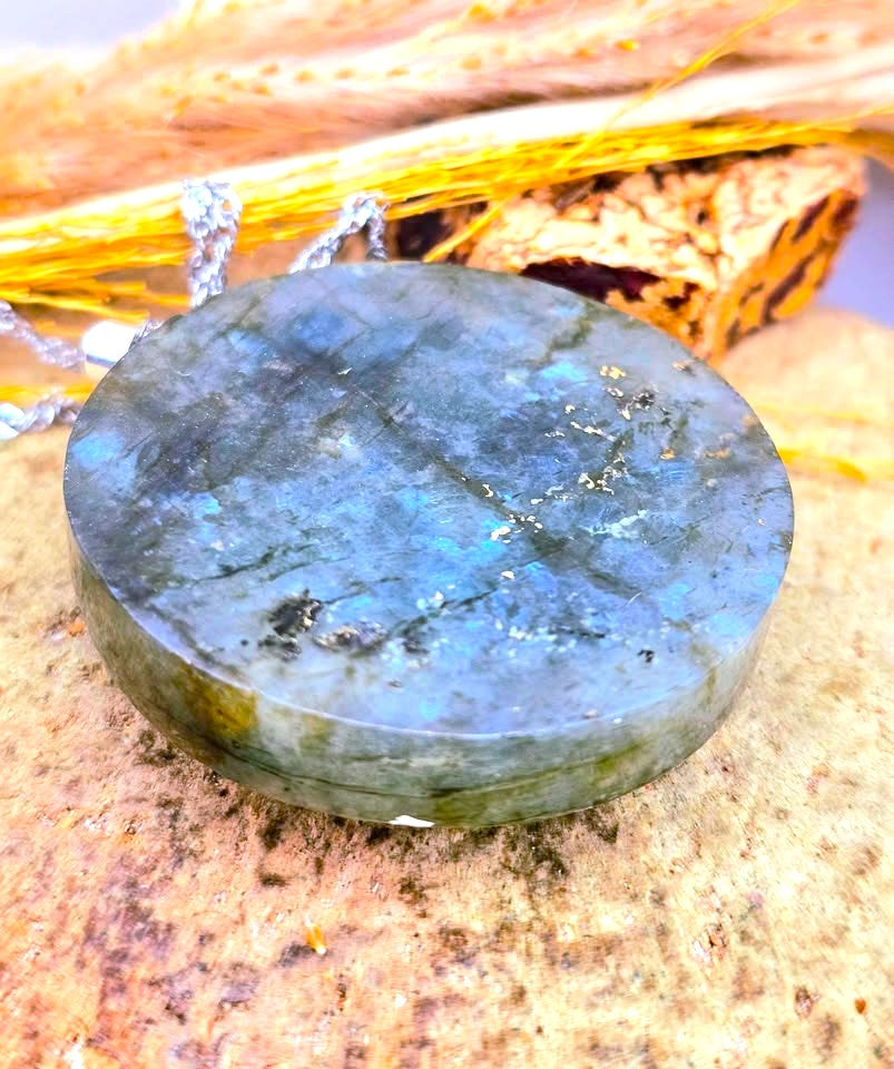 A polished round labradorite gemstone pendant with a silver chain, resting on a textured stone surface with dried grass in the background.