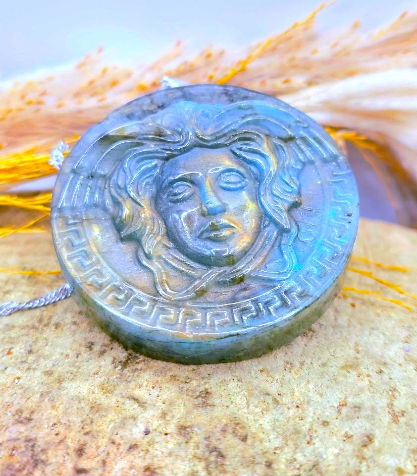 A round labradorite pendant featuring a raised Medusa head design and Greek key border, resting on a textured stone with dried grass in the background.