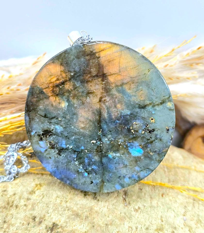 A round labradorite stone pendant with a silver bail, resting on a textured stone surface with dried grass in the background.