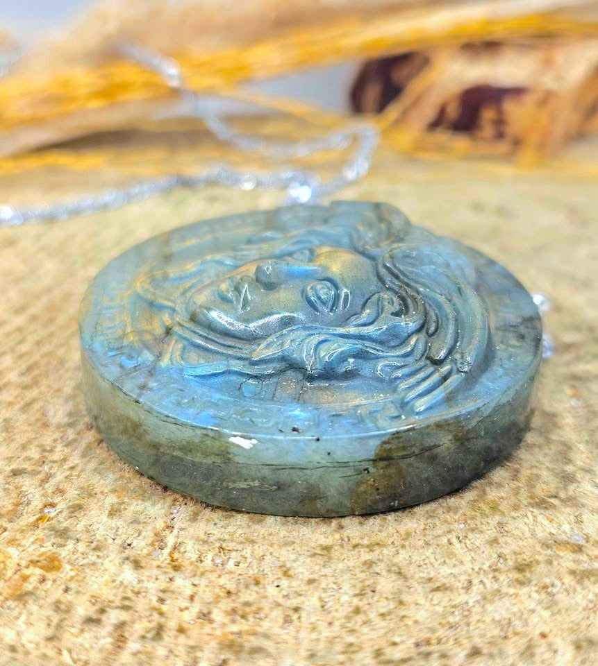 A round carved labradorite pendant necklace featuring a detailed face design, resting on a textured tan surface with a silver chain visible in the background.