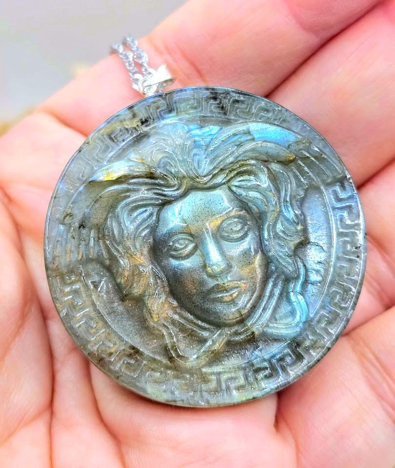 A round labradorite pendant featuring a Medusa-style face relief and Greek key border, held in a person's hand. The pendant has a silver bail with a chain, and the stone displays a blue-green iridescent labradorite play-of-color.
