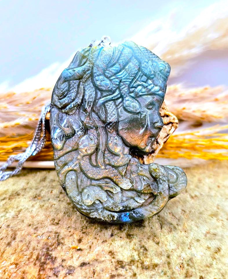 A carved labradorite pendant featuring a profile of a woman's face surrounded by swirling wave-like patterns, resting on a textured rock with a blurred natural background.
