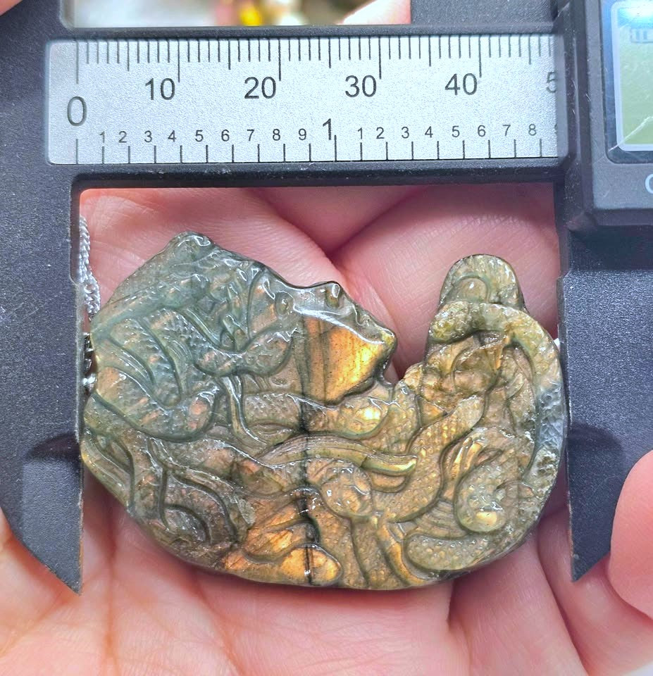 A carved stone pendant with an iridescent gold and green finish, held in a person's hand under a digital caliper to show its dimensions.