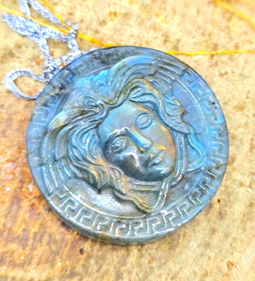 A round pendant necklace featuring a sculpted Medusa head design with a Greek key border, crafted from a metallic blue-gray material, hanging from a silver chain on a textured stone surface.