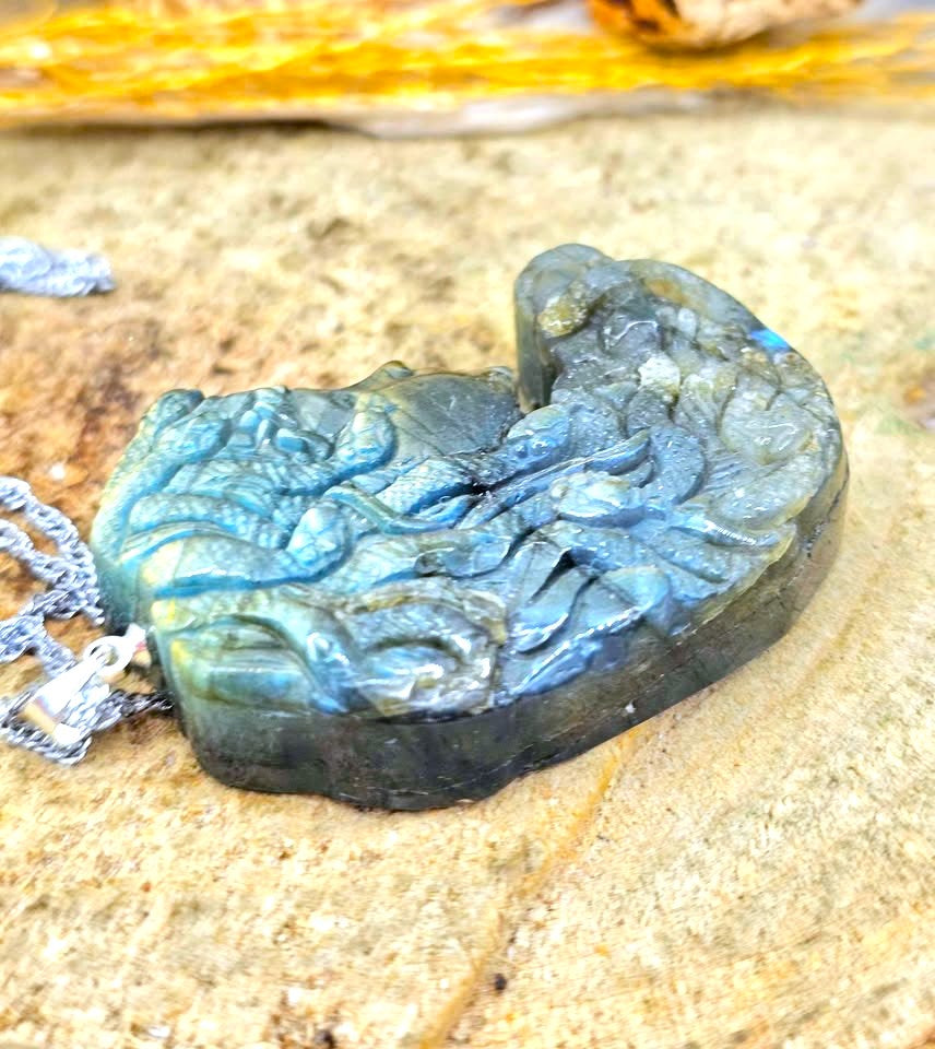 A carved labradorite pendant with a detailed design, resting on a textured stone surface with a silver chain partially visible.