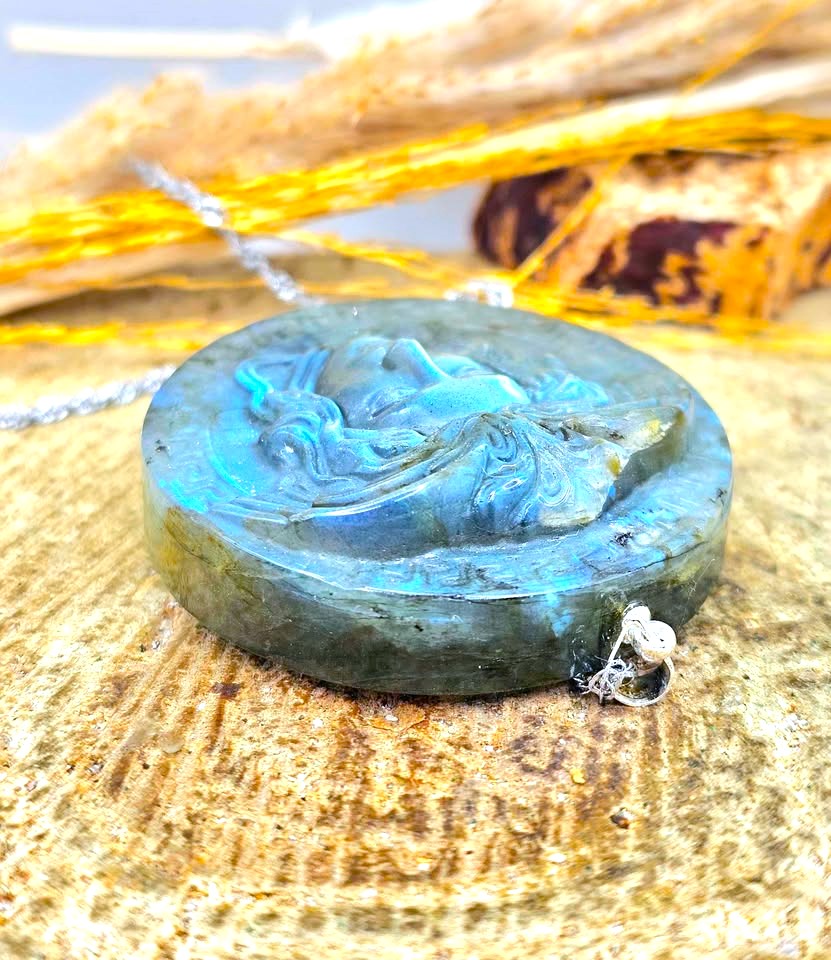 A round carved labradorite pendant with a silver bail, resting on a textured wooden surface with dried grass in the background.