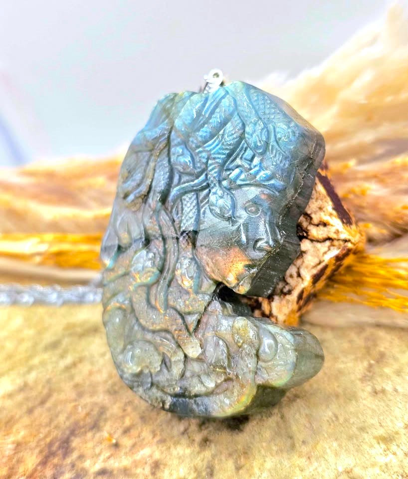 A carved labradorite pendant featuring a detailed mermaid design, resting on a piece of driftwood with a blurred natural background.