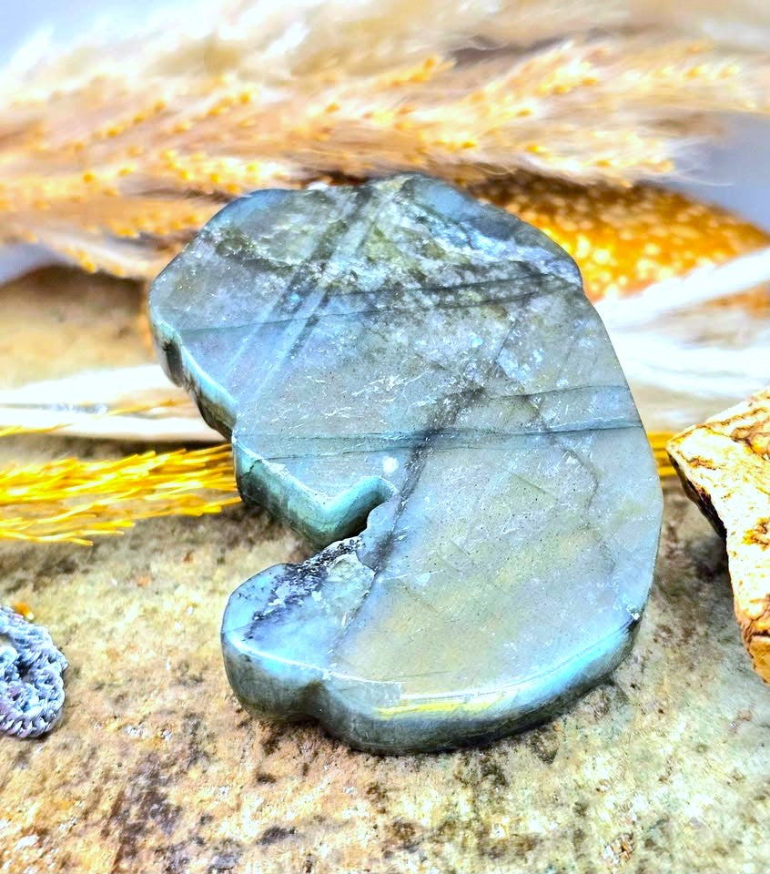 A polished Labradorite pendant with a natural organic shape, resting on a textured stone surface with decorative feathers in the background.