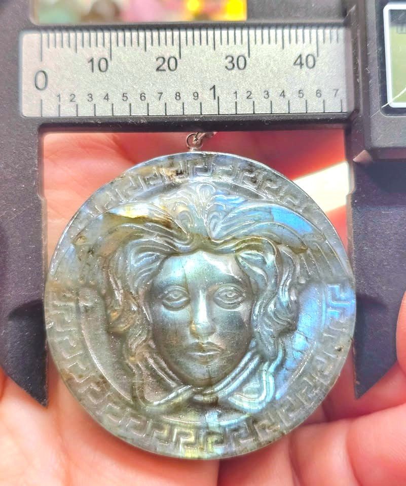 A round labradorite pendant featuring an embossed Medusa head design, held in a hand under a digital caliper with a centimeter ruler scale visible above it.