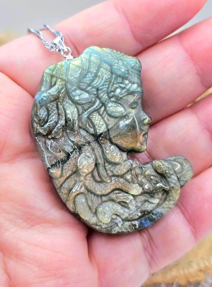 A hand holding a carved labradorite pendant necklace featuring a profile of a woman surrounded by snakes, with a silver bail.