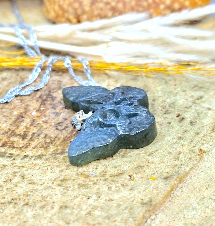 A dark gray, textured stone pendant shaped like a four-petal flower, hanging from a silver-toned chain on a sandy surface.