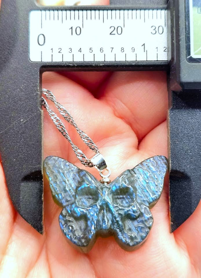A butterfly-shaped pendant necklace with a textured, iridescent blue-gray finish, held in a person's hand under a metal caliper to show its size. The pendant is attached to a silver chain.