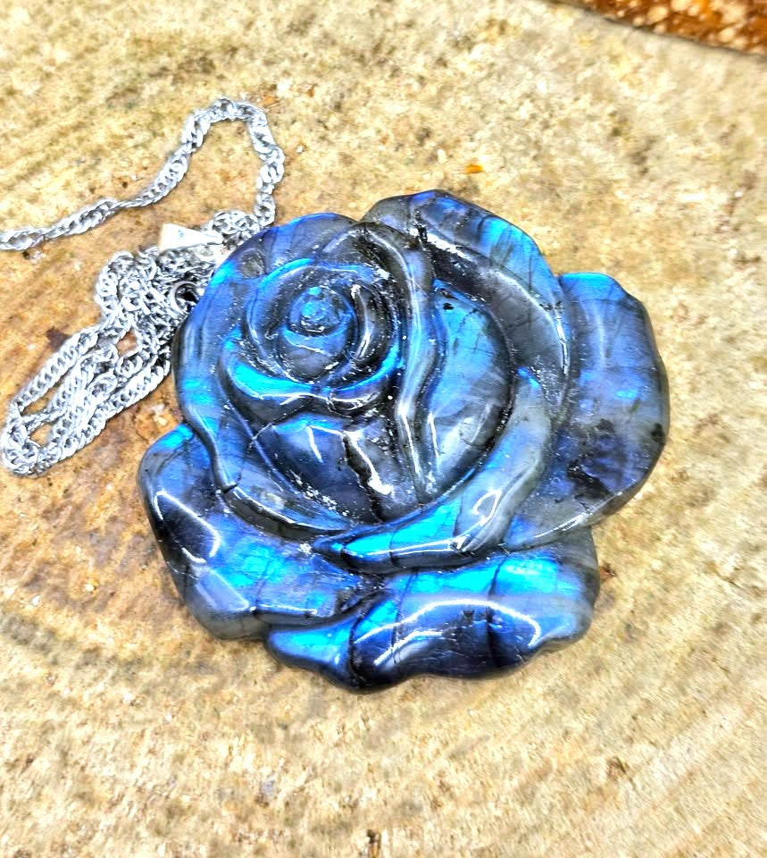 A carved rose-shaped pendant made of iridescent blue labradorite stone, shown with a silver chain on a textured tan stone surface.