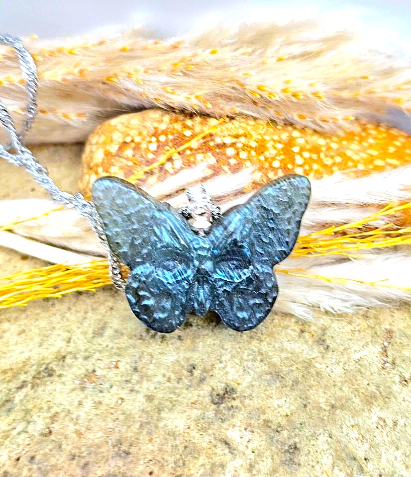 A blue iridescent butterfly pendant necklace with a silver-toned setting and chain, displayed on a textured stone surface with dried grass and yellow string in the background.