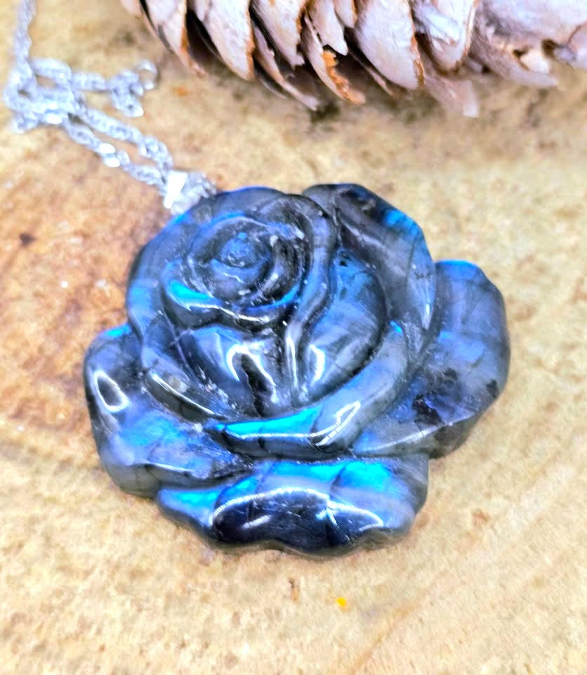 A polished dark blue and black rose-shaped pendant necklace with iridescent blue flashes, hanging from a silver chain on a textured tan surface with a pinecone in the background.