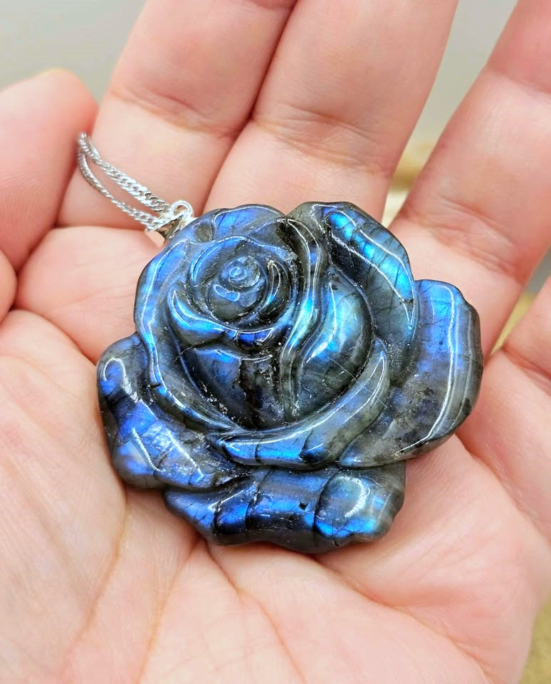 A close-up of a hand holding a carved labradorite rose pendant on a silver chain. The pendant displays a vibrant blue and green iridescent labradorite effect with dark gray stone showing through.