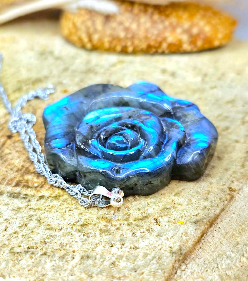 A close-up of a carved rose-shaped pendant necklace made from iridescent labradorite, showing blue and green flashes, attached to a silver chain on a sandy surface.
