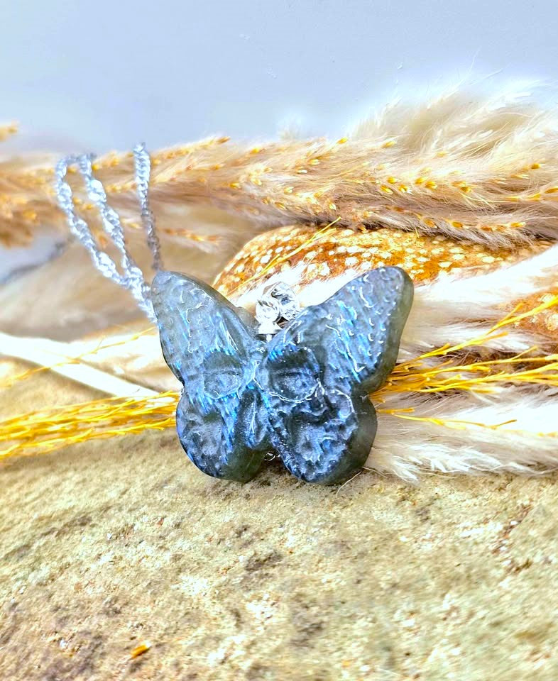 A blue butterfly-shaped pendant necklace with a textured, stone-like surface, resting on a sandy surface with dried grass and a small silver clasp.