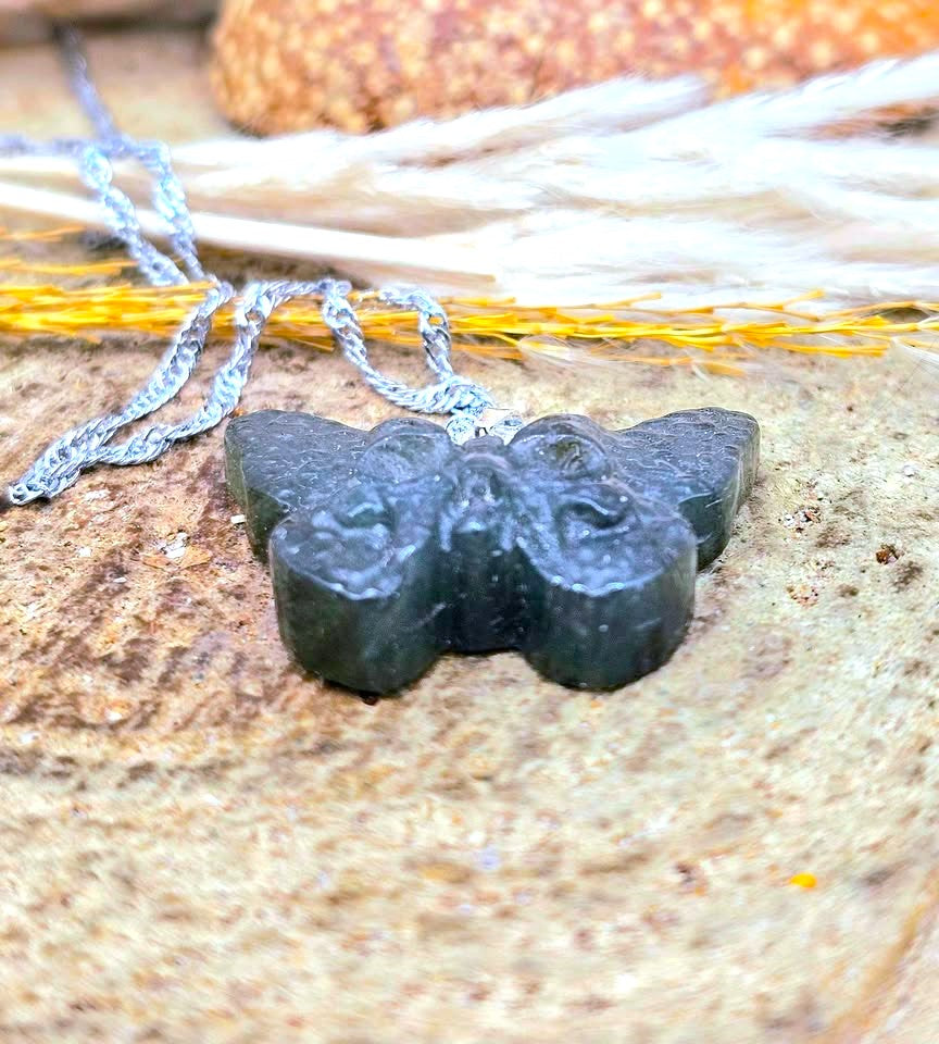 A close-up of a dark carved stone pendant on a silver chain, resting on a textured stone surface with dried grass and a small orange object blurred in the background.
