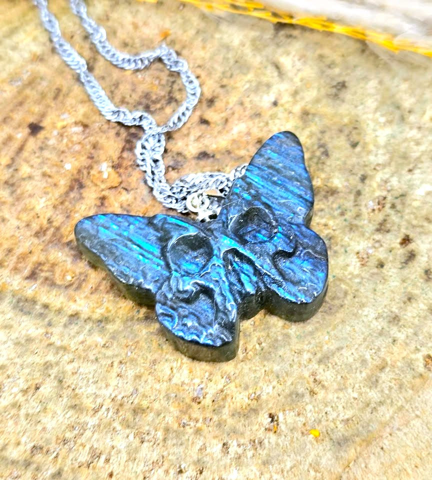 A silver chain necklace with a butterfly-shaped pendant carved with a skull design, featuring a blue iridescent finish, resting on a textured stone surface.
