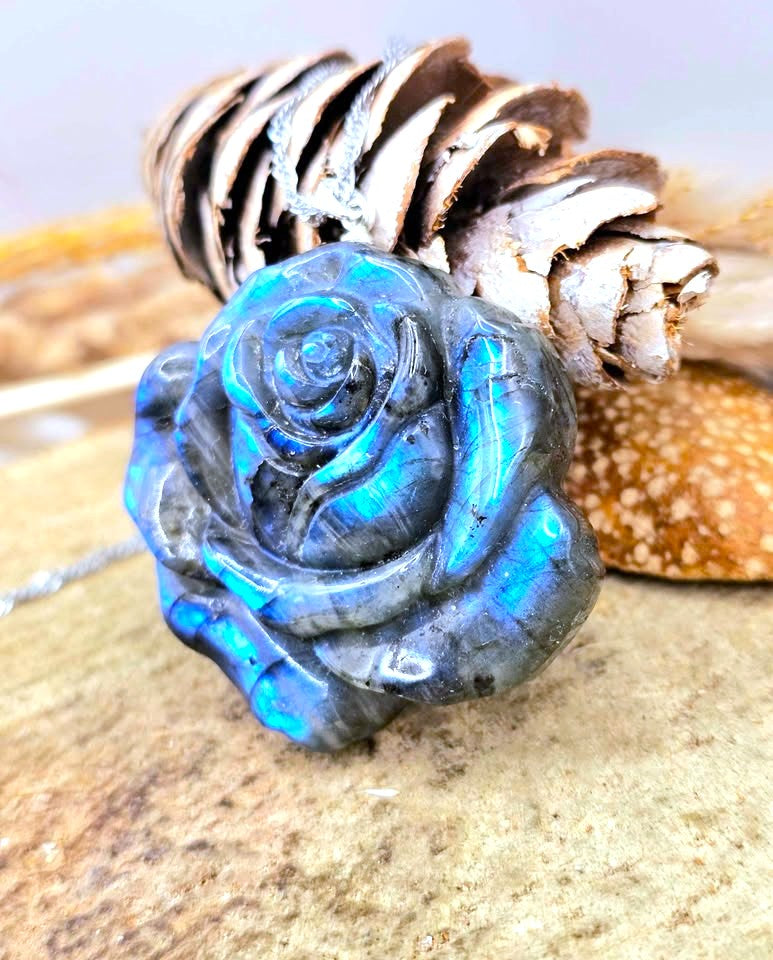 A carved labradorite rose pendant with a vibrant blue iridescent sheen, displayed on a textured stone surface with a pinecone and seashell in the background.