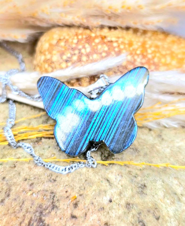 A butterfly-shaped pendant necklace with a glossy blue and black striped pattern, resting on a textured stone surface with a silver chain.